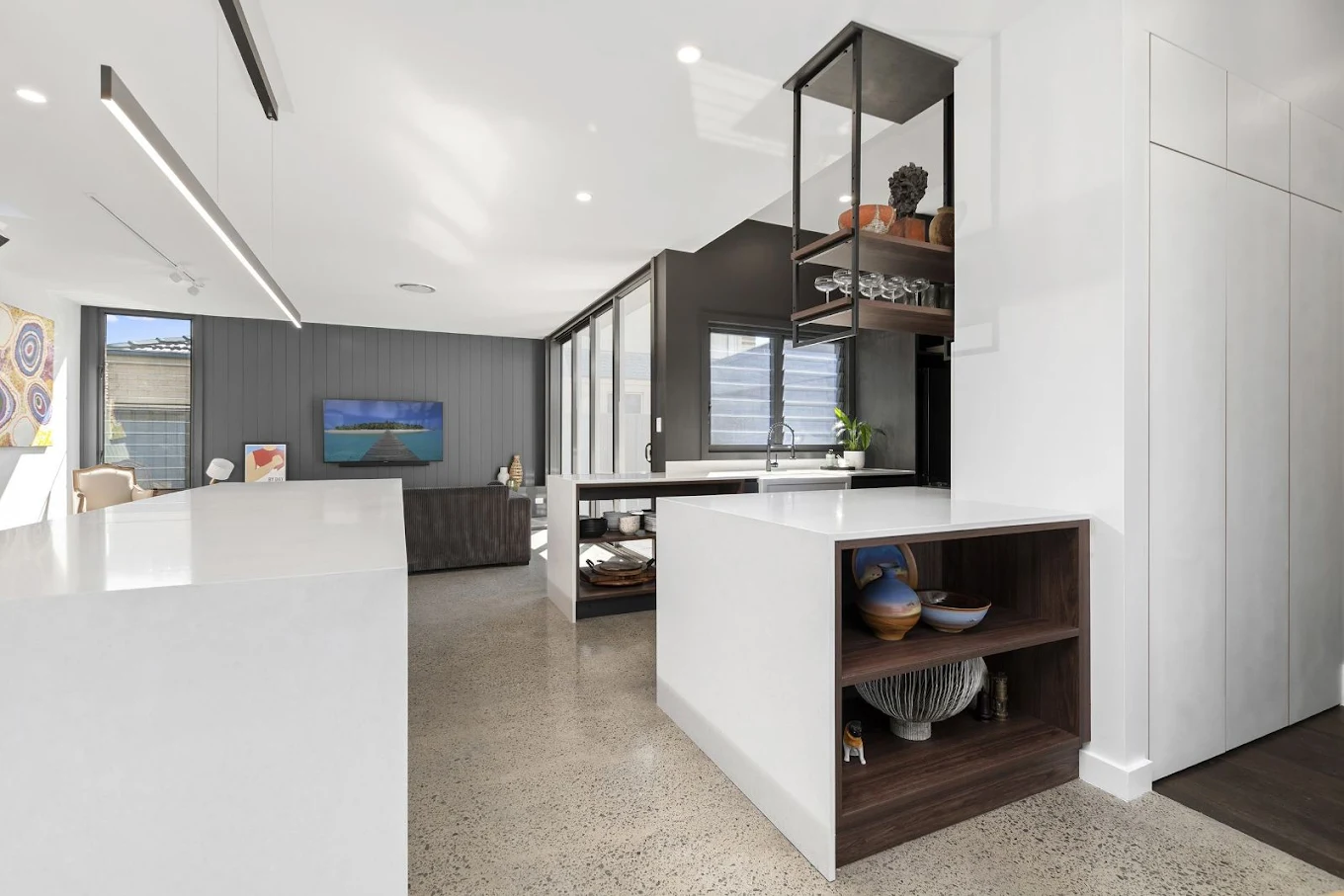 Open-plan kitchen and living area with white island bench, suspended metal shelving, walnut accents and polished concrete floor by MTS Group