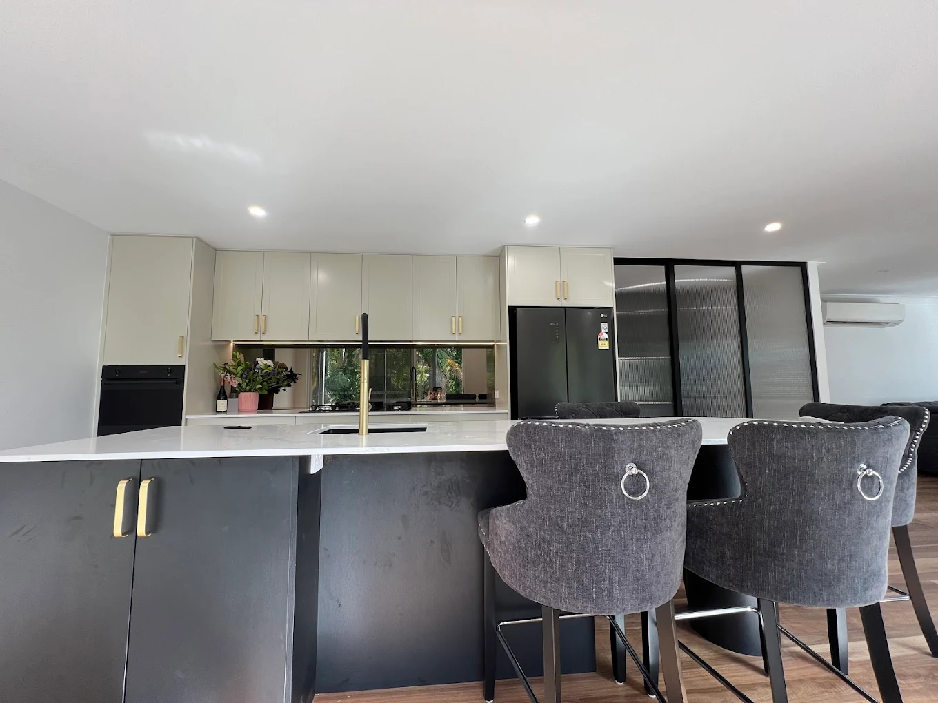 Kitchen renovation with mirror splashback, brass tapware, dark island bench with upholstered bar stools by MTS Group