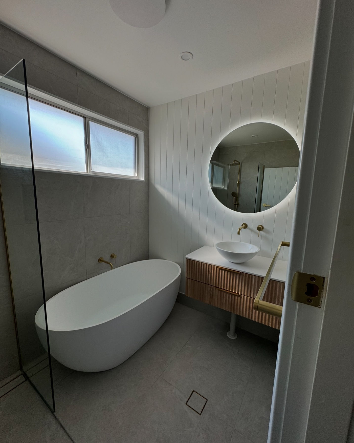 Bathroom renovation with freestanding bath, fluted timber vanity, LED-backlit round mirror and brass fixtures by MTS Group
